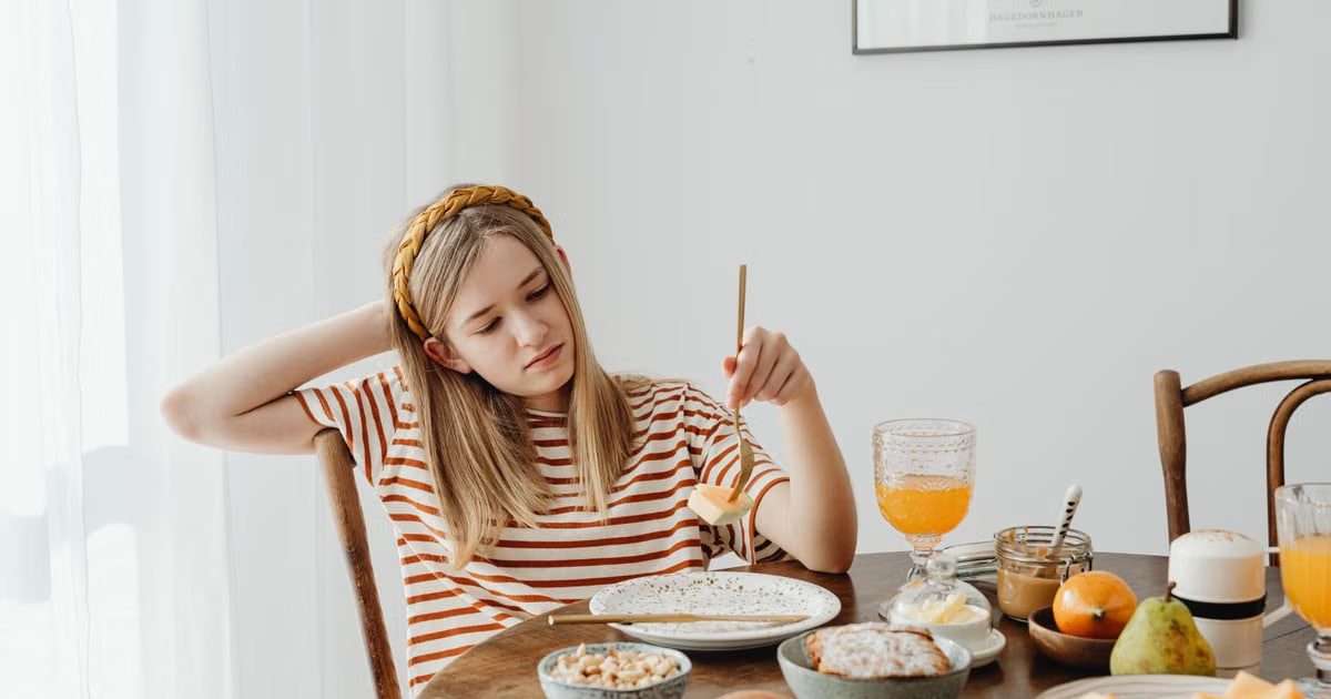 Teen girl in striped shirt at breakfast table with fruits, bread, and juice. Casual morning setting.
